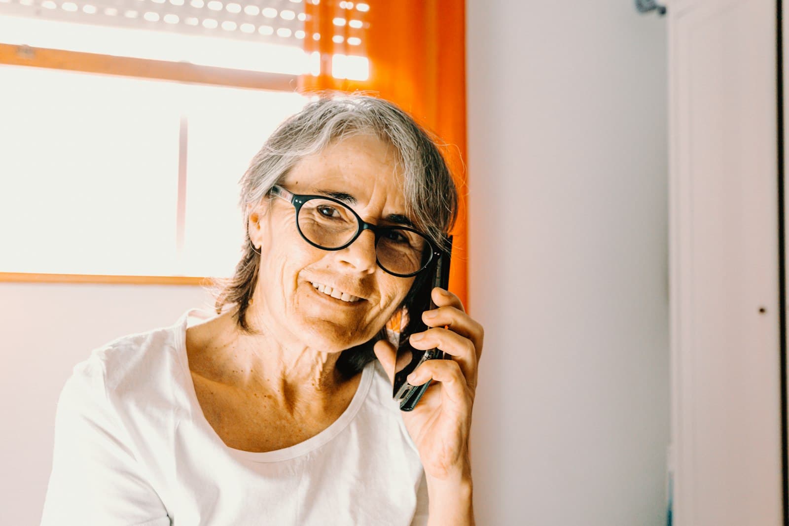 An older woman with glasses smiling while talking on the phone at home